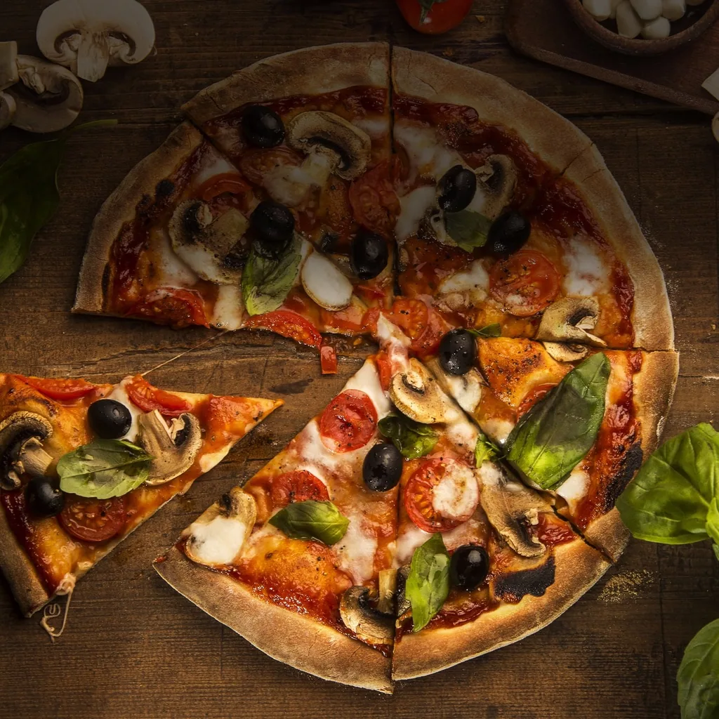 Knusprige Steinofen-Pizza von oben auf rustikalem Holzbrett mit Champignons, Oliven, Tomaten und Basilikumblättern.