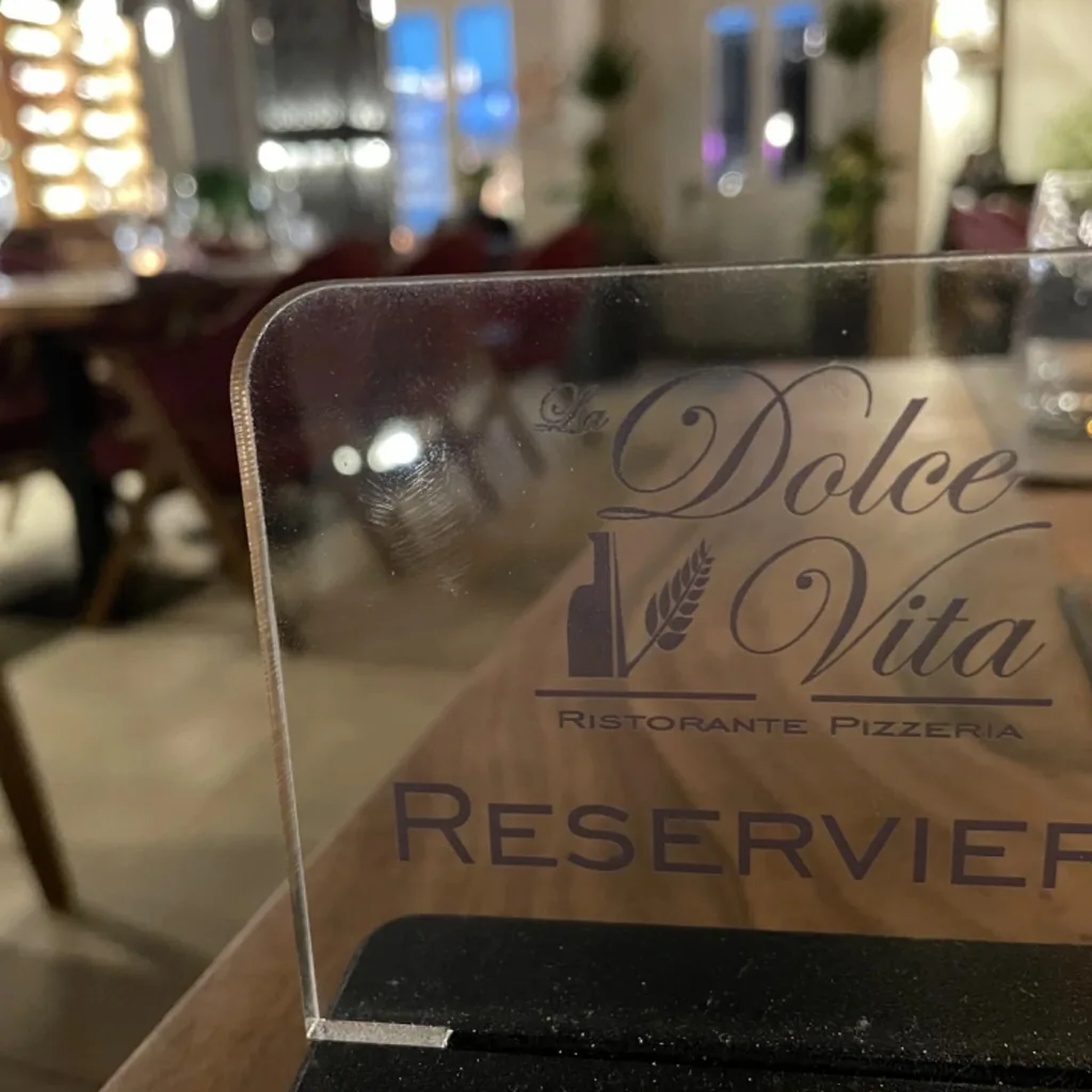 Graviertes Acrylschild «La Dolce Vita Ristorante Pizzeria Reserviert» auf massivem Holztisch mit Gastraum im Hintergrund.
